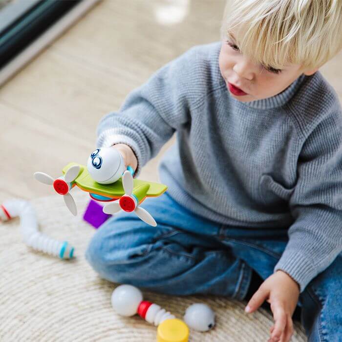 Child holding a green and white magnetic toy with propellers, with colorful building pieces on a rug.