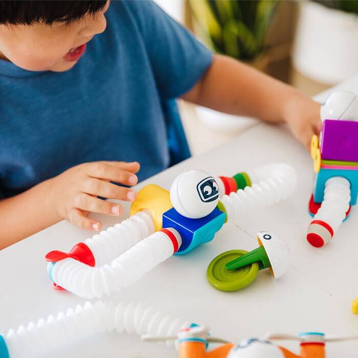 Child building colorful robots using flexible white tubes, magnetic blocks, and spheres with robot faces.