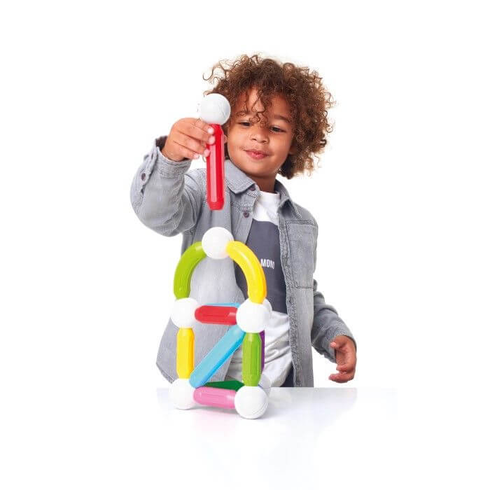 A young child builds a structure using colorful red, yellow, and green magnetic rods and white spheres.