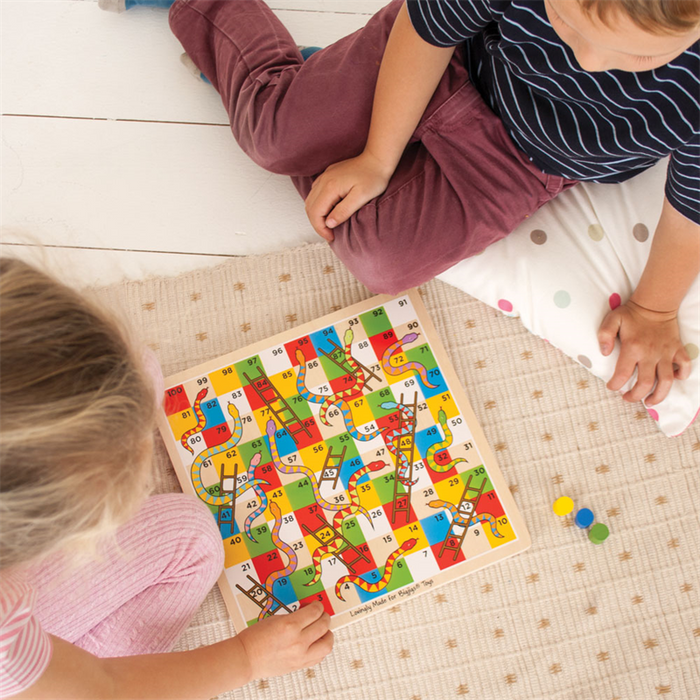 Two children playing a colorful wooden board game with numbered squares, snakes, and ladders on a light rug.