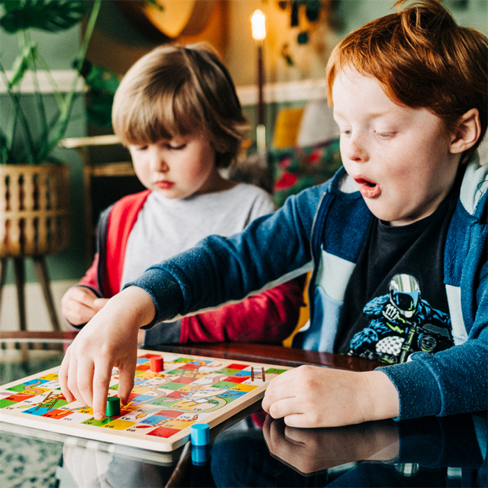 Two young boys playing a wooden Snakes and Ladders board game with colorful graphics and cylindrical playing pieces.