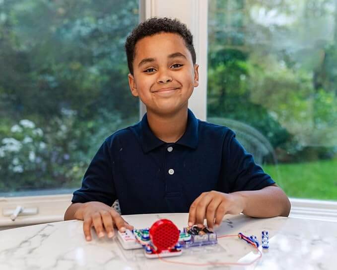 Smiling boy assembles Snap Circuits FM Radio with a red speaker and colorful components on a white marble table.