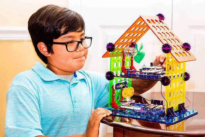 A boy with glasses looks at a Snap Circuits house with an orange roof, green walls, blue base, and visible electronics.