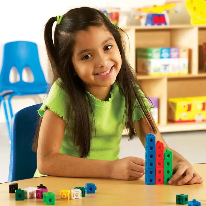 A smiling girl builds towers using colorful, interlocking plastic cubes on a classroom table.