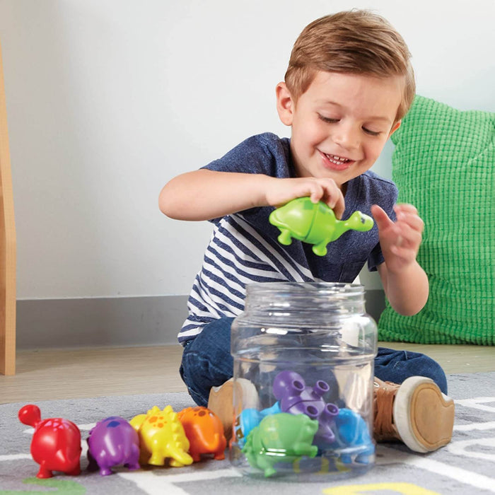 A child drops a green dinosaur into a clear jar. Colorful snap-together dinosaurs are lined up on a rug.