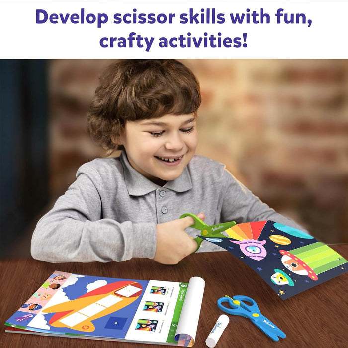 Smiling child cuts space-themed paper with green safety scissors next to a craft book, blue scissors, and a glue stick.