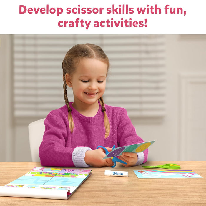 Smiling girl uses blue safety scissors to cut colorful paper activities near a craft book and glue stick.