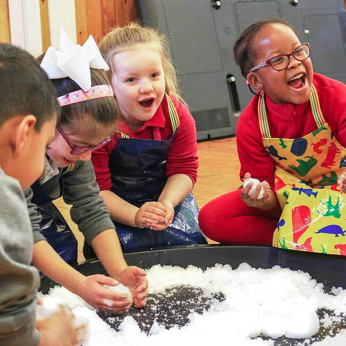 Four children excitedly shape snowballs from white artificial snow in a large black tray on a wooden floor.