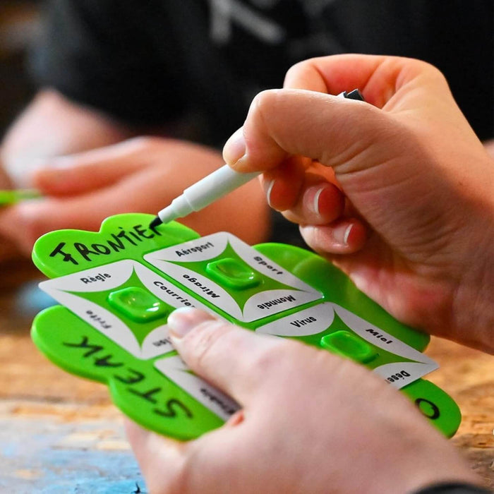 A person writing on a green clover-shaped game board with word cards slotted in using a dry-erase marker.