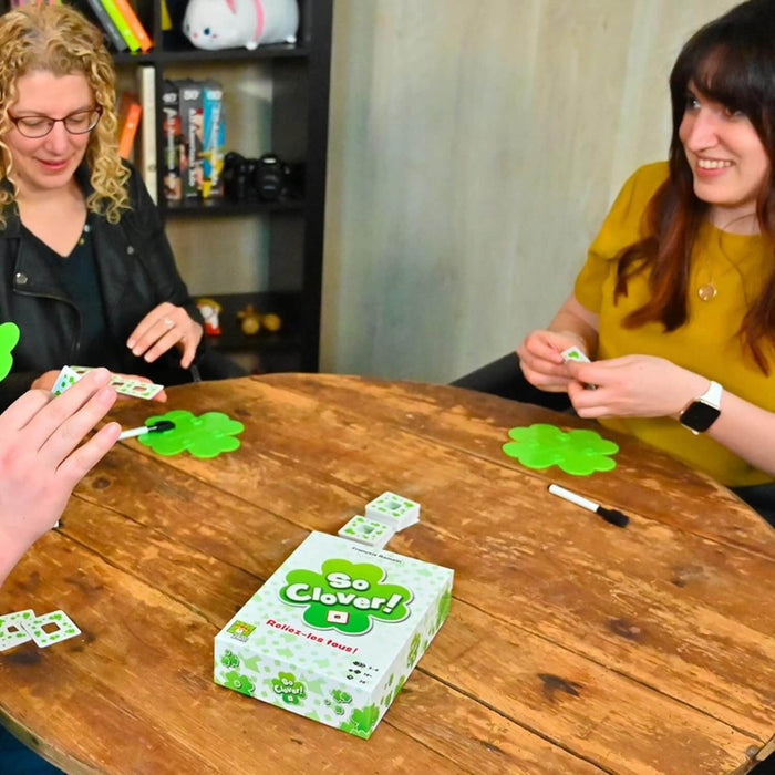 People playing So Clover! featuring a white box with green clovers, clover-shaped boards, and cards on a wooden table.