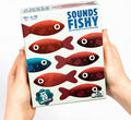 Hands hold a board game box featuring red fish and one blue fish swimming in opposite directions on a white background.