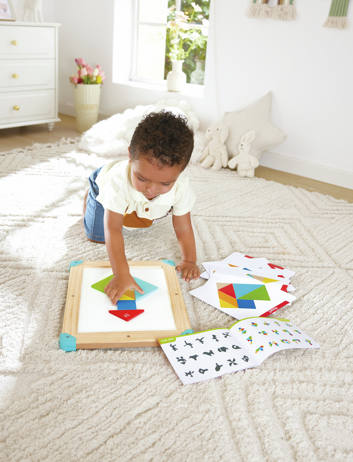 Toddler using colorful magnetic geometric shapes on a wooden tangram board with pattern cards and a guide booklet.