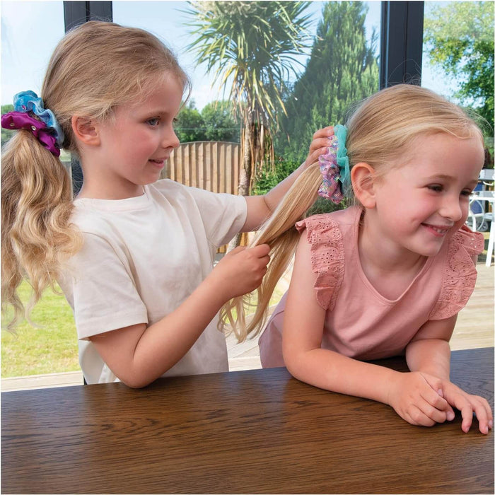 Two girls styling long blonde hair with sparkly, sequined scrunchies in pink, teal, and purple.