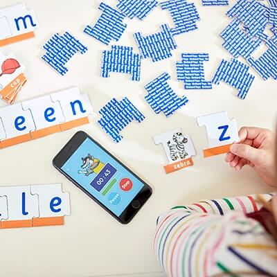 Child matches blue letter puzzle pieces and a zebra card next to a smartphone timer app on a white table.