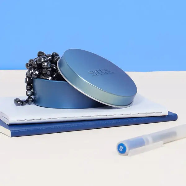 Shiny black ferrite magnets spilling from a round blue metal tin on a stack of notebooks.