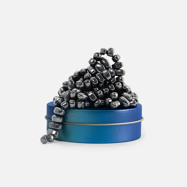 A heap of gunmetal grey magnetic stones on a blue-to-teal gradient metal tin against a white background.