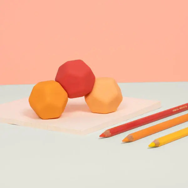 Three red and orange faceted silicone shapes stacked on a pink surface beside three matching colored pencils.
