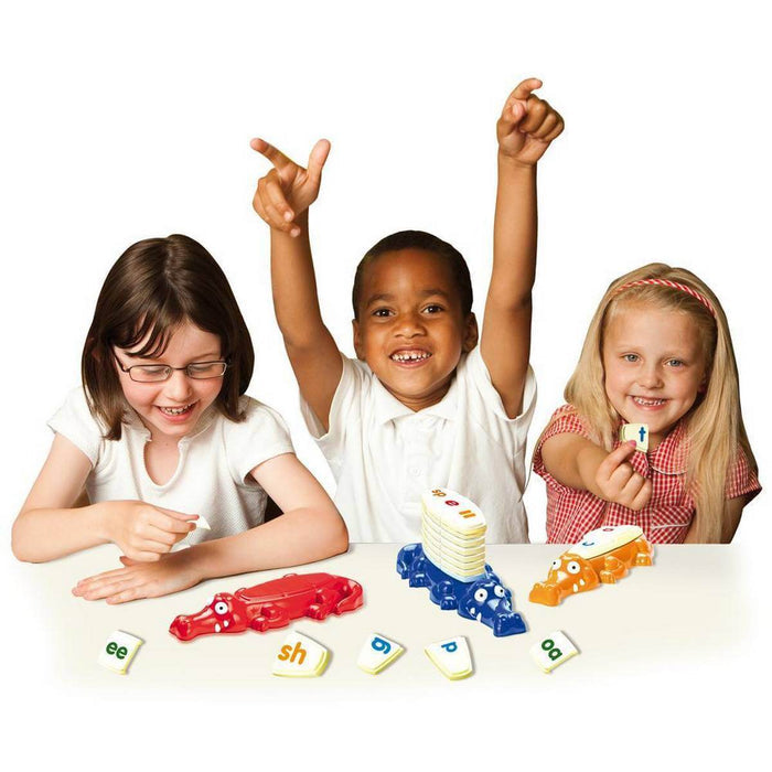 Three children playing with red, blue, and orange crocodile-shaped tile holders and white letter tiles.