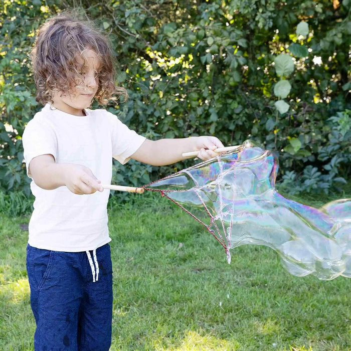 Child creates a massive, iridescent bubble using wooden sticks and a red string spiderweb wand outdoors.