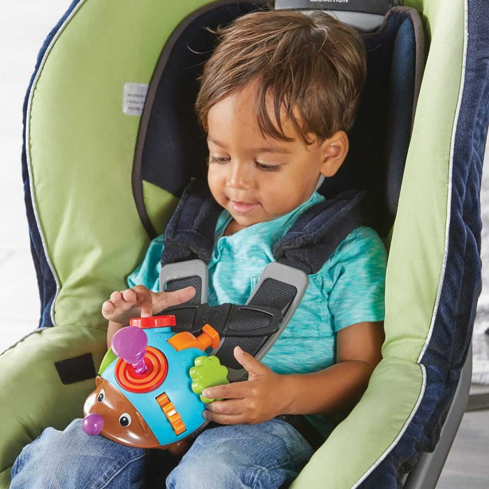 Toddler in a car seat playing with a blue and brown hedgehog toy with colorful tactile knobs, dials, and gears.