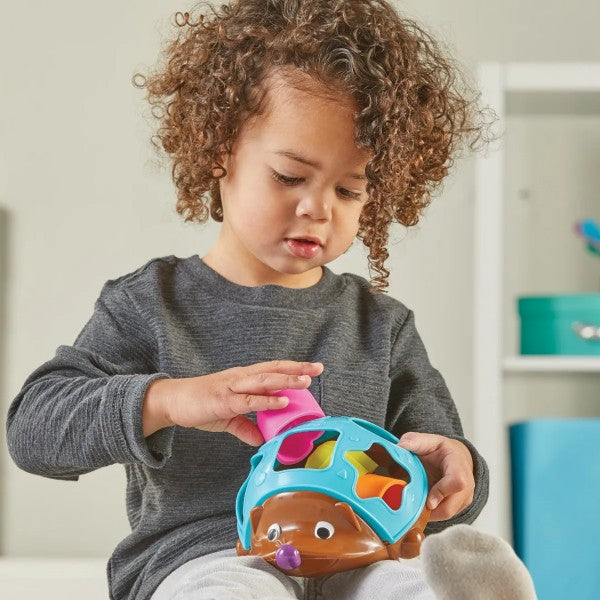 Curly-haired child inserting a pink block into a blue and brown hedgehog shape sorter toy.