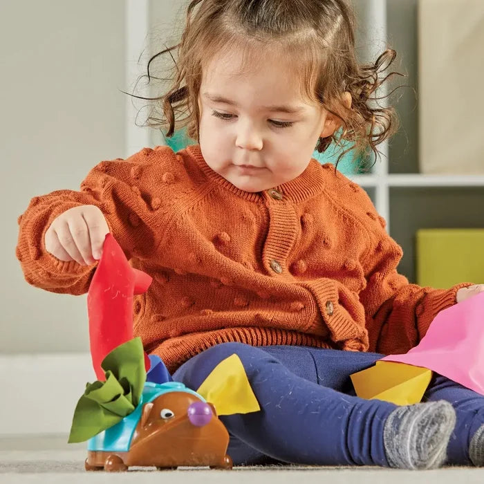Toddler pulls colorful fabric squares from a brown and teal hedgehog toy with a purple nose.