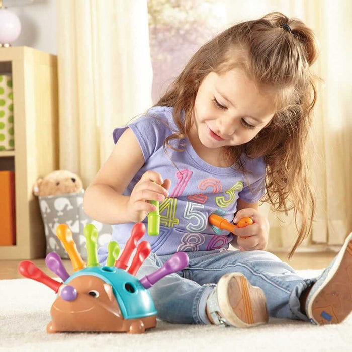 A smiling girl inserts colorful plastic pegs into a teal and brown hedgehog toy while sitting on a rug.