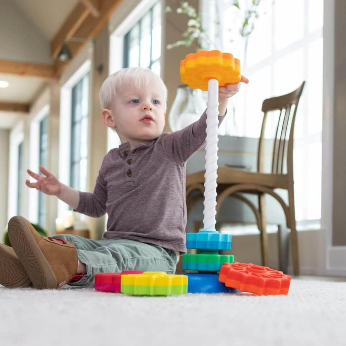 Toddler stacking colorful gear-shaped discs onto a white spiral pole toy while sitting on a white rug.