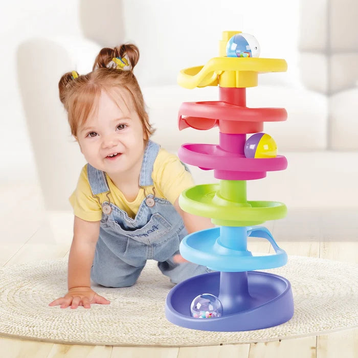 Baby girl crawling next to a six-tier rainbow spiral ramp toy with colorful balls on a light rug.