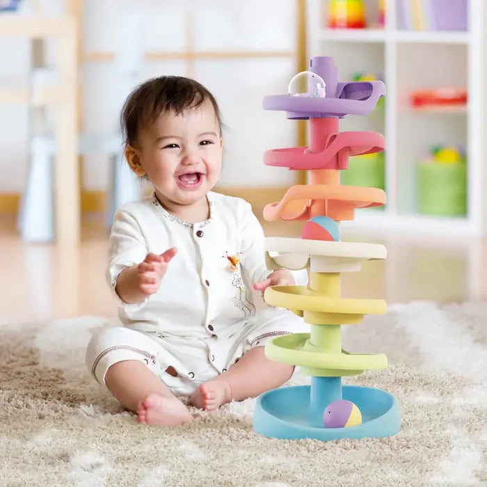 Smiling baby sitting by a seven-tier pastel spiral ball tower with rolling balls on a fluffy rug.