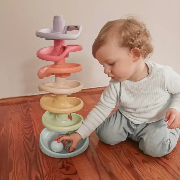 Toddler playing with a 7-tier spiral ball ramp made of muted pastel recycled plastic discs on a wooden floor.