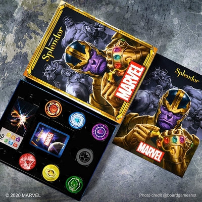 Splendor Marvel board game box with Thanos cover art and open tray of colorful symbol tokens on a grey stone surface.