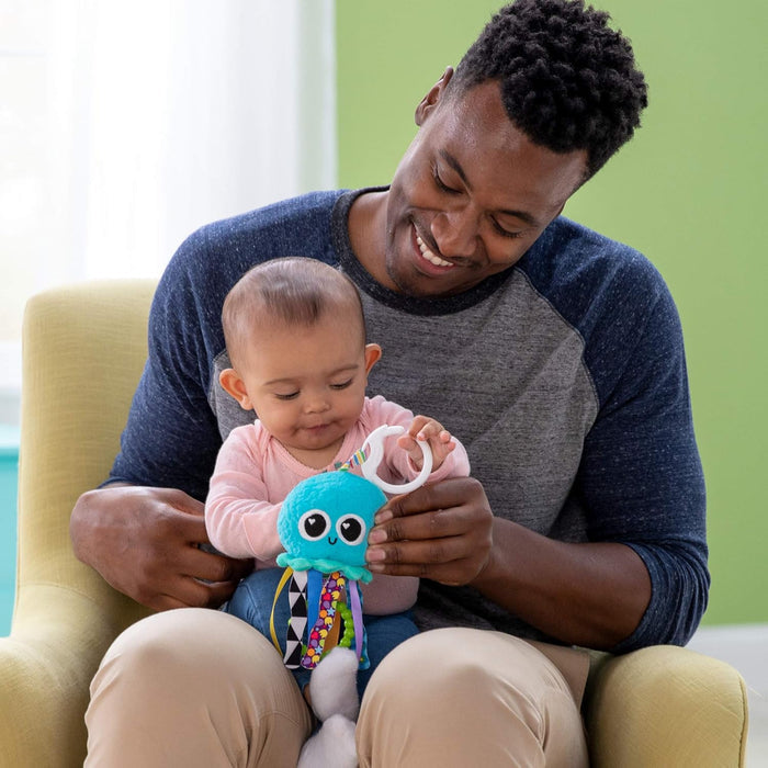 Man and baby play with a blue plush jellyfish toy featuring colorful ribbon tentacles and a white plastic clip.