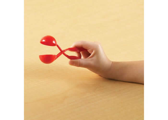 Child's hand using red scissor-style tweezers with round scoops on a light wood background.