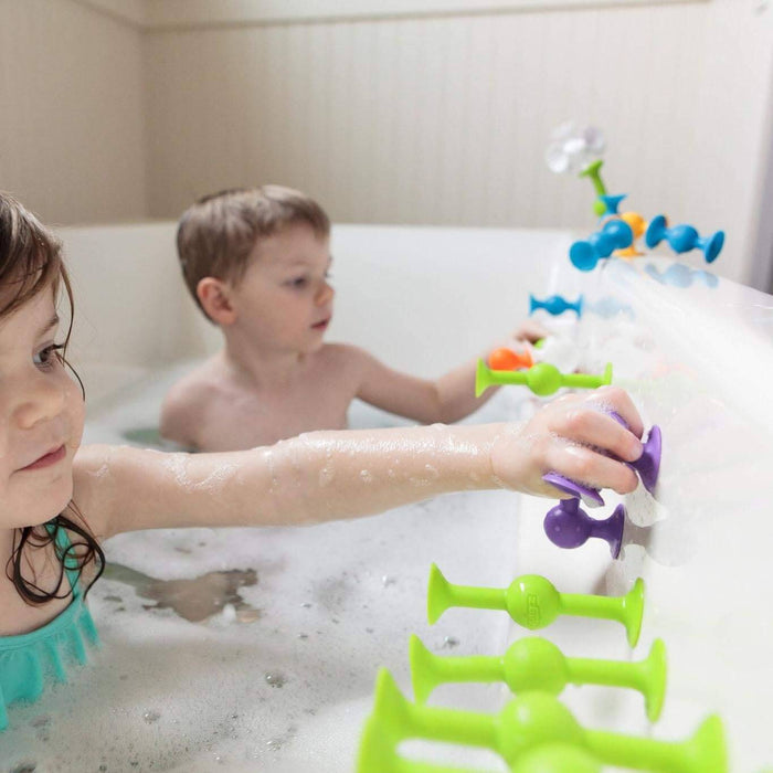 Two kids in a bubble bath stick colorful green, blue, and purple suction cup toys to the side of the white bathtub.