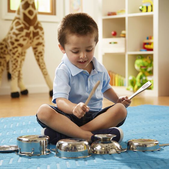 A boy plays on a blue rug with shiny stainless steel toy pots, pans, and a colander using a wooden spoon and spatula.