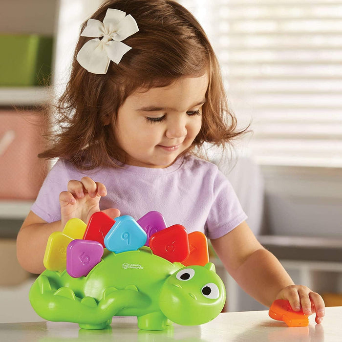A toddler playing with a green dinosaur toy by inserting colorful, numbered plates into slots on its back.