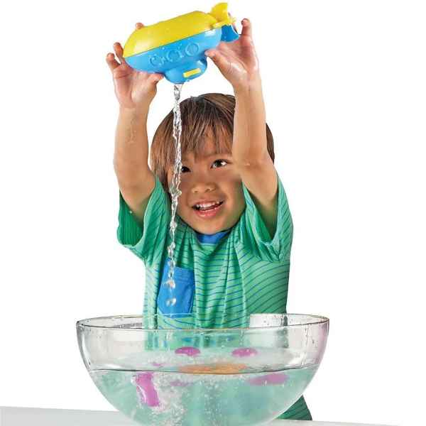Child pours water from a blue and yellow toy submarine into a clear bowl with floating purple and orange shapes.