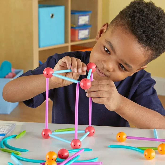 A young boy builds a 3D geometric shape using pink and blue rods with red connector spheres on a white table.