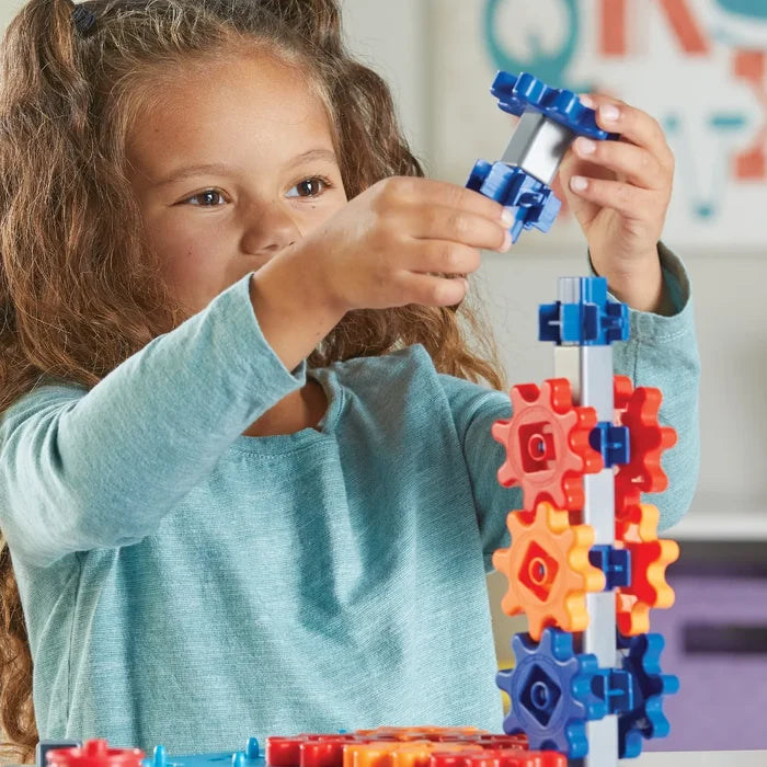 Young girl building a tower by stacking colorful blue, red, and orange toy gears onto a vertical post.