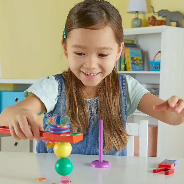 Smiling girl lifts colorful magnetic discs with an orange wand balanced on stacked yellow and green balls.