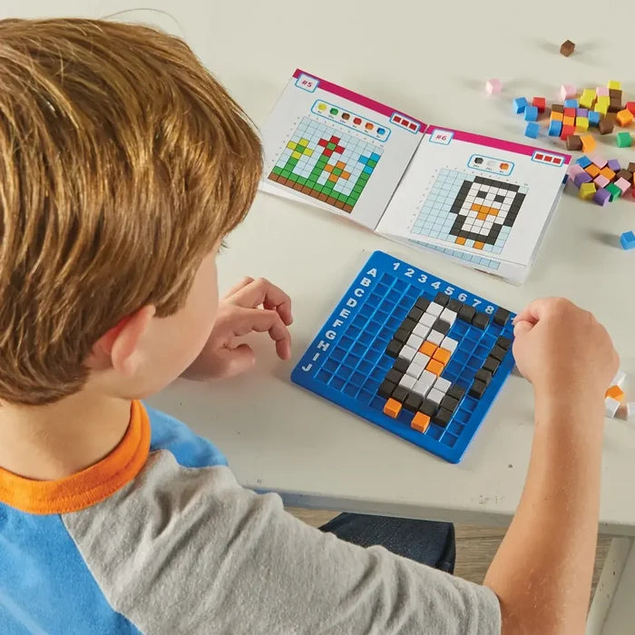 Child builds a pixel penguin on a blue grid board using colored cubes, following an instruction booklet.