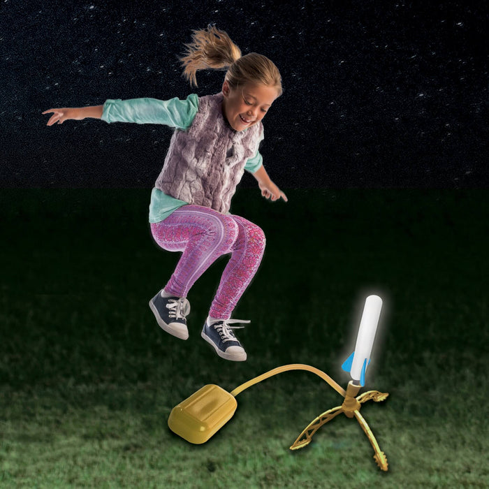 Child jumps to stomp a yellow pump, launching a glowing white foam rocket with blue fins into the starry night sky.