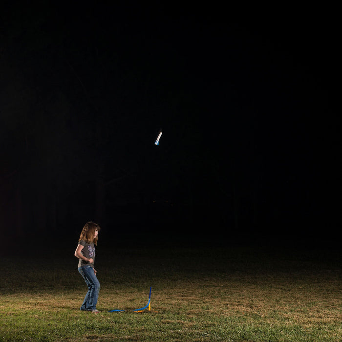 A young girl launches a glowing white toy rocket into the night sky from a blue stomp launcher in a grassy field.
