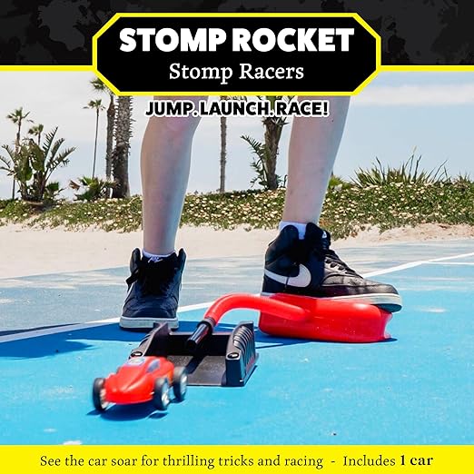 Person stomps on a red air pump to launch a red toy car from a black ramp onto a blue outdoor court near a beach.