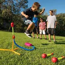 Boy jumps on pink air pad to launch red-tipped foam rocket from a yellow stand with blue tubing on a grassy field.