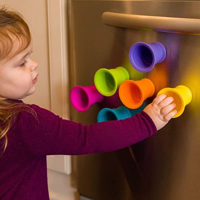 Toddler reaching for colorful, funnel-shaped silicone toys suctioned to a stainless steel appliance.