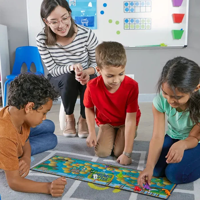 A teacher and three children play a colorful swamp-themed board game with lily pad paths on a gray classroom rug.