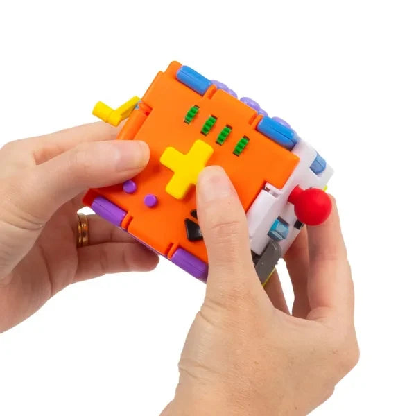 Hands holding a vibrant orange sensory cube featuring a yellow D-pad, green wheels, and a red joystick.