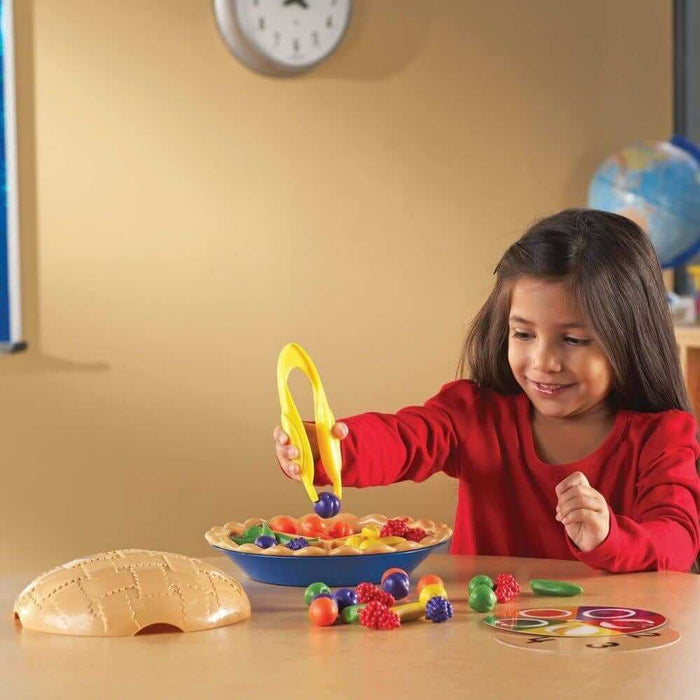 Girl uses yellow tweezers to sort colorful fruit pieces into a blue toy pie dish with a tan crust lid nearby.